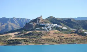 Pueblo blanco en la cima de una colina rocosa, con un castillo en ruinas. Un lugar ideal que ver cerca de Ronda, con un embalse azul en primer plano.
