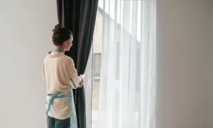 Kobieta w fartuszku odsłania zasłony, odsłaniając białe firanki. Zastanawia się, co ile zabki w karniszu, by idealnie je zawiesić.