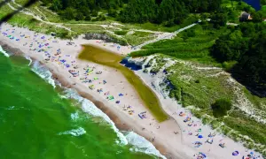 Plaża w Bobolinie tętni życiem. Ludzie odpoczywają na piasku, kąpią się w morzu i spacerują. W oddali widać las i malowniczą wydmę.
