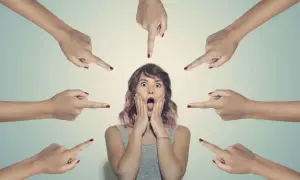 Una ragazza con espressione spaventata, circondata da dita puntate. La sua bocca è spalancata, le mani sulle guance, simbolo della paura del giudizio altrui.