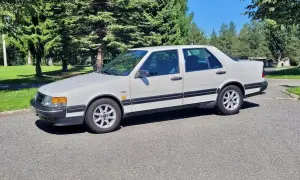 Vit Saab 9000 CD parkerad på en asfalterad väg med gröna träd och en blå himmel i bakgrunden.