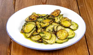 Piatto di zucchine alla poverella, fette sottili e dorate condite con olio e erbe aromatiche, su un piatto bianco con bordo blu.