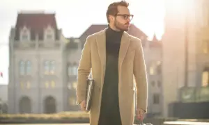 Mann im eleganten Mantel und Rollkragenpullover, mit Laptop und Koffer, verkörpert den "old money style" vor historischer Kulisse.