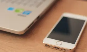 Biały smartfon leży obok laptopa. W tle widać logo z kolorowymi ikonami, sugerującymi uruchomione aplikacje w tle.