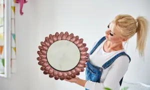 Okrągłe lustro w ramie z metalowych łyżeczek, jak zrobić je samemu. Kobieta w ogrodniczkach prezentuje efekt DIY.