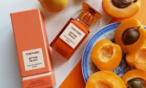 Pomarańczowe opakowanie i buteleczka perfum Tom Ford Bitter Peach otoczone soczystymi brzoskwiniami.