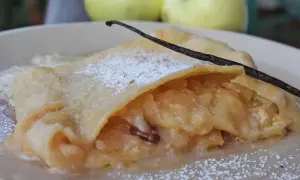 Ein Stück bayerischer Apfelstrudel, bestäubt mit Puderzucker und garniert mit einer Vanilleschote, daneben zwei Äpfel.