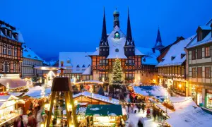 Ein verschneiter Weihnachtsmarkt in Deutschland mit beleuchteten Buden, einem Weihnachtsbaum und einem historischen Rathaus im Hintergrund.