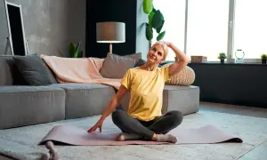 Donna bionda fa stretching al collo, esercitando l'elevatore della scapola, seduta su un tappetino in soggiorno.