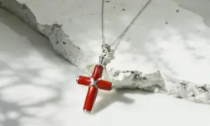 Collar con colgante de cruz de coral rojo y plata, sobre una superficie texturizada de piedra blanca.