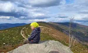 Dziecko w żółtej czapce siedzi na skale, patrząc na górzysty krajobraz z lasami i szlakiem.