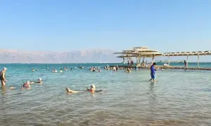 Ludzie unoszą się na wodzie Morza Martwego, korzystając z jego niezwykłego zasolenia. W tle góry i plaża z zacienionymi miejscami.