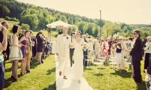 Szczęśliwa para młoda idzie alejką podczas ślubu w lipcu, otoczona gośćmi na tle zielonych wzgórz.
