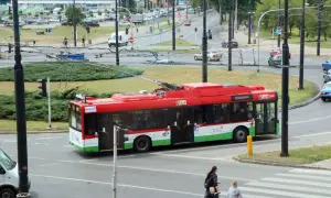 Czerwony trolejbus z napisem "Lublin" przejeżdża przez skrzyżowanie. W tle widać ruch uliczny i budynki.
