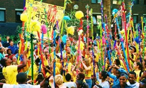 Gente celebra en un desfile, levantando coloridos bastones adornados con pompones y cintas. Un cartel dice "POLE-O-NG STATION". Parece una fiesta, quizás un bank holiday.