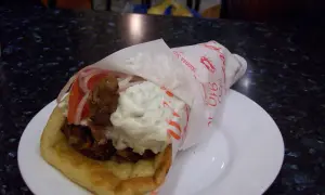 Pyszna pita grecka z mięsem, warzywami i sosami, zawinięta w papier z greckimi napisami.