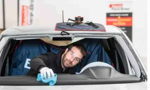 Un technicien porte des lunettes de protection et utilise un chiffon bleu pour nettoyer l'intérieur du pare-brise d'une voiture.
