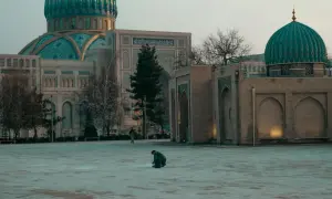 Samotny podróżnik klęczy na pustym placu przed meczetem z turkusową kopułą.