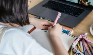 Dziewczynka pisze w zeszycie, obok leży laptop i piórnik z kredkami.