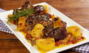 Un saporito agnello con patate, cotto lentamente con pomodorini e rosmarino, servito su un piatto bianco.