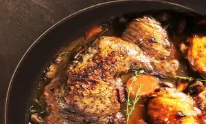 Un plat de coq au vin mijoté, avec des morceaux de poulet dorés, des carottes, des champignons et des herbes dans une sauce riche.