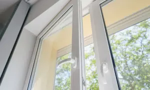 Białe okno z profili pvc, uchylone, z widokiem na zielone drzewa.