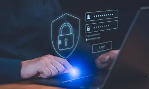 Dłoń dotyka ekranu laptopa z interfejsem logowania. Ikona tarczy z kłódką symbolizuje bezpieczeństwo, a obok pola na login i hasło znajduje się przycisk "Login". To nowoczesny menedżer haseł.