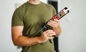 Mężczyzna w zielonej koszulce trzyma pistolet z tubą kleju do listew przypodłogowych plastikowych.