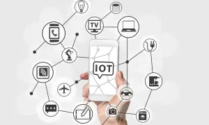 Dłoń trzyma smartfon z napisem "IOT co to". Wokół widnieją ikony urządzeń połączonych siecią: żarówka, TV, laptop, ekspres do kawy, pralka, samochód, aparat, samolot, robot, smartfon, router i chmura danych.