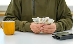 Osoba liczy banknoty 200 zł, symbolizujące podwyżki dla pracowników samorządowych. Obok stoi żółty kubek i portfel.