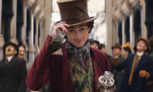 Timothée Chalamet w filmach, jako młody Willy Wonka, w cylindrze i barwnym stroju, z uśmiechem pozdrawia tłum.
