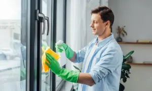 Mężczyzna w zielonych rękawiczkach czyści okna plastikowe, używając sprayu i żółtej ściereczki. Dbałość o detale to klucz do konserwacji okien.