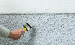 En hand målar en putsad fasad med en pensel. Det är en del av huset som är målad vit och en del som är grå.