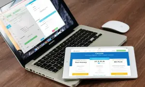 Programista .NET pracuje nad aplikacją na laptopie i tablecie. Na ekranach widoczne są formularze i cennik usług.
