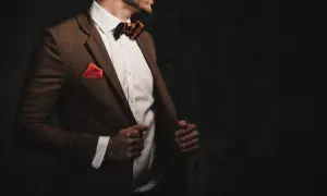 Elegancki mężczyzna w brązowej, tweedowej marynarce w kratkę, białej koszuli i muszce. Stylizacje z tweedową marynarką to klasyka.