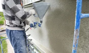 Pracownik nakłada tynk silikonowy natryskowo na elewację budynku za pomocą agregatu.