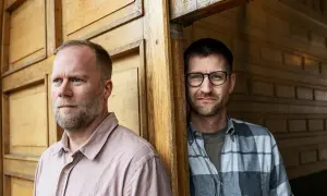 Två män, en med skägg och ljus skjorta, den andra med glasögon och rutig skjorta, står vid en trävägg. Mikael Wolf är en av dem.