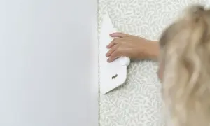 En hand använder en vit spackel för att släta ut en tapetsera non woven med ett ljust bladmotiv.