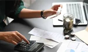 Persona calcola spese per indennità trasferta con calcolatrice e fogli. Barattolo pieno di monete e laptop sullo sfondo.