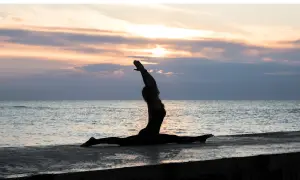 Silhouette di una donna in Ardha Hanumanasana sulla riva del mare al tramonto, con le braccia alzate verso il cielo.