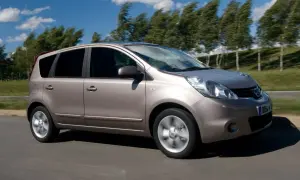 Złoty Nissan Note 2010 pędzi po drodze, drzewa na tle falują na wietrze.