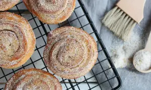 Nybakade kanelbullar med strössel av brunt socker och pärlsocker på ett svart galler. En träpensel och en sked med socker ligger bredvid.
