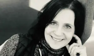 Karin Ståhl ler mot kameran, med handen vid kinden. Hon bär hörlurar runt halsen och en väst över en mönstrad tröja.