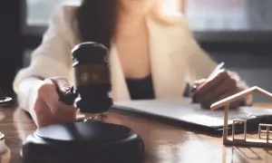 Sądowy młotek i model domu symbolizują licytację komorniczą. Kobieta podpisuje dokumenty, przygotowując się do sprzedaży nieruchomości.