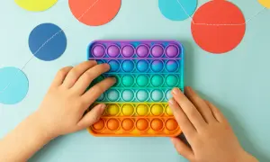 Dziecięce dłonie bawią się tęczowym pop it na niebieskim tle z kolorowymi kółkami.