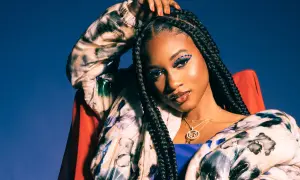 Nija Charles, with striking blue eye makeup and braids, poses confidently in a colorful jacket against a blue backdrop.