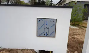 Metalowa skrzynka gazowa z napisem "GAZ" zamontowana na białym, otynkowanym murze.