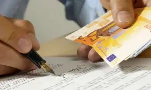 Ein Mann unterschreibt ein Dokument mit Füllfederhalter, während er Geldscheine hält. Dies könnte ein Beispiel für einen zahlung unter vorbehalt musterbrief sein.