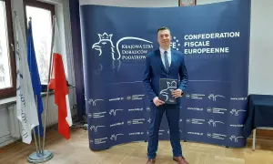 Mężczyzna w garniturze stoi przed niebieskim banerem z logo Krajowej Izby Doradców Podatkowych.