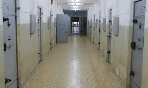 Korytarz więzienny z rzędami cel. Drzwi cel, niektóre z numerami jak 106 i 222, są zamknięte. W głębi widać kraty i otwarte drzwi.